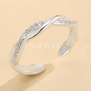 Puede incluir: Un anillo plateado con un diseño retorcido. Un lado del anillo está incrustado con pequeñas piedras transparentes. El anillo abierto en la parte inferior presenta el texto "LN JEWELS" y "S925".
