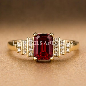May include: A gold ring featuring a large, rectangular, deep red gemstone as the centerpiece. The ring is accented with smaller, clear gemstones on each side of the main stone. The band is a polished gold color.