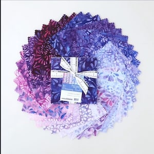 May include: A stack of square fabric swatches arranged in a circular fan pattern, featuring a gradient of purple, violet, and lavender floral batik prints. A white ribbon with ruler markings secures the center, labeled Dancing Petals.