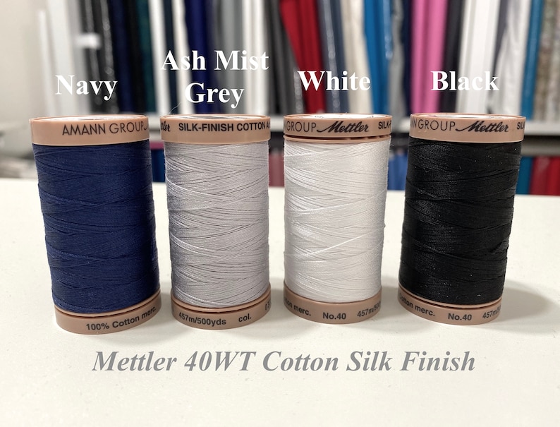 Może przedstawiać: Cztery szpulki nici Mettler 40WT Cotton Silk Finish w kolorze granatowym, popielatym, białym i czarnym. Każda szpulka jest oznaczona logo Mettler oraz wagą i rodzajem nici.