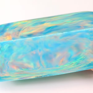 May include: A rectangular block of resin with a smooth, semi-translucent finish. The object features a swirling pattern of bright sky blue, soft white, and iridescent gold hues that create a shimmering, light-reflecting effect across the surface.