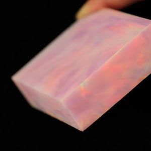 May include: A faceted, rectangular gemstone with a translucent appearance. The stone displays a blend of pink, orange, and purple hues, with a subtle internal glow. The gemstone is held against a black background.