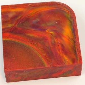 May include: A rectangular block with rounded corners, showcasing a vibrant blend of colors. The surface features swirling patterns of red, orange, yellow, and hints of purple, creating a marbled effect. The block appears to be made of a solid, possibly resin, material.