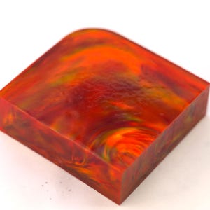 Op de afbeelding: Een vierkant blok met afgeronde hoeken, met een wervelend patroon van rode, oranje en gele tinten. De randen zijn dieprood, wat een levendig, gemarmerd effect creëert. Afmetingen niet zichtbaar.