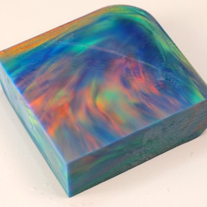 Op de afbeelding: Een vierkant, doorschijnend blok met afgeronde hoeken. Het blok heeft een wervelend patroon van blauwe, groene, oranje en rode kleuren, wat een levendige uitstraling creëert. Het object is waarschijnlijk een decoratief item of een kunstwerk.
