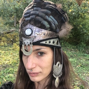 May include: A headdress featuring dark feathers, a silver and black band with a central white stone, and dangling silver ornaments. The headpiece has a tribal or bohemian aesthetic, suitable for festivals or themed events.