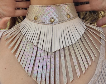 Può includere: Un'ampia collana girocollo caratterizzata da una fascia con texture a pelle di serpente iridescente e borchie in bronzo metallizzato. Il pezzo è decorato con due strati di lunghe frange in pelle color crema che pendono a V, creando un effetto stratificato.