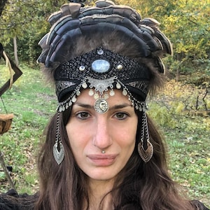 May include: A black and silver headpiece with feathers, beads, and dangling charms. The headpiece features a central oval stone and a sun-shaped pendant. The background is a natural outdoor setting with trees and foliage.