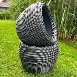 Puede incluir: Dos cestas de mimbre gris tejidas apiladas sobre hierba verde. Las cestas tienen forma redondeada y un interior oscuro. El patrón de tejido es visible, creando una superficie texturizada. El fondo presenta plantas verdes altas y una casa.