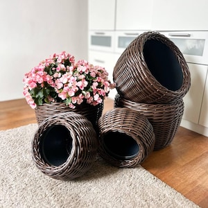 Puede incluir: Un conjunto de cuatro maceteros de mimbre trenzado en color marrón oscuro con revestimientos interiores negros lisos. Un macetero contiene un grupo de pequeñas flores rosas con hojas verdes, mientras que los otros tres están vacíos y apilados.