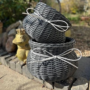May include: Two dark gray woven baskets stacked vertically, each featuring a rounded shape and a smooth texture. A thin, white decorative ribbon is tied in a bow around the center of each basket, providing a contrast against the dark material.
