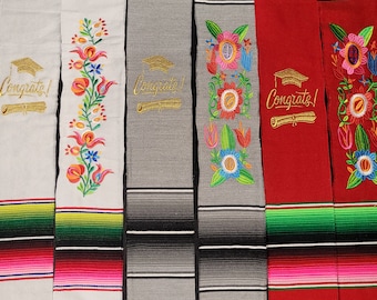 May include: Four graduation sashes with colorful stripes and embroidered flowers. The sashes are white, gray, red, and blue. Each sash has the word "Congrats!" embroidered in gold with a graduation cap and scroll.