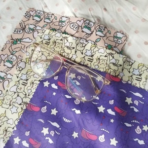 May include: A pair of clear glasses with a gold frame on top of three patterned microfiber cloths. The cloths have a variety of cartoon characters and designs, including owls, stars, and a purple creature with a tongue sticking out.