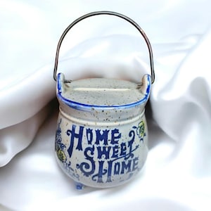 Puede incluir: Una alcancía de cerámica blanca con forma de olla y un asa de metal. Tiene diseños florales azules y amarillos y la frase "Home Sweet Home" escrita en ella.