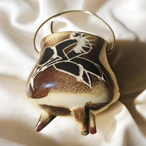 Puede incluir: Un pequeño caldero de cerámica con un esmalte marrón y crema, con un diseño floral en blanco y negro. Tiene tres patas pequeñas y un asa de metal plateado. El caldero probablemente se utiliza con fines decorativos o rituales.