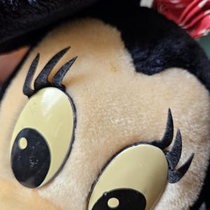 May include: Close-up of a plush toy mouse with black fur, large black eyes, and long black eyelashes. The toy has a beige face and a red bow.