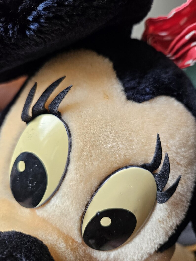 May include: Close-up of a plush toy mouse with black fur, large black eyes, and long black eyelashes. The toy has a beige face and a red bow.