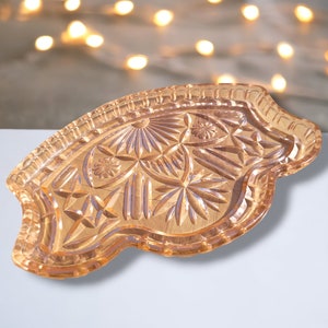 Könnte beinhalten: Ein pfirsichfarbenes, dekoratives Glastablett mit aufwendigen Schnitzmustern. Das Tablett hat einen gewellten Rand und eine Vielzahl von geometrischen und floralen Designs. Das Tablett wird wahrscheinlich zum Servieren oder Ausstellen verwendet.