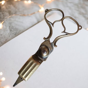 May include: Antique brass scissors with a unique heart-shaped design. The scissors have a pointed tip and a decorative handle.