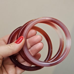 May include: Three translucent bangles in shades of red and pink are held in a hand. The bangles have a smooth, glossy finish and vary in color intensity, from deep burgundy to pale pink. The background is a neutral beige.