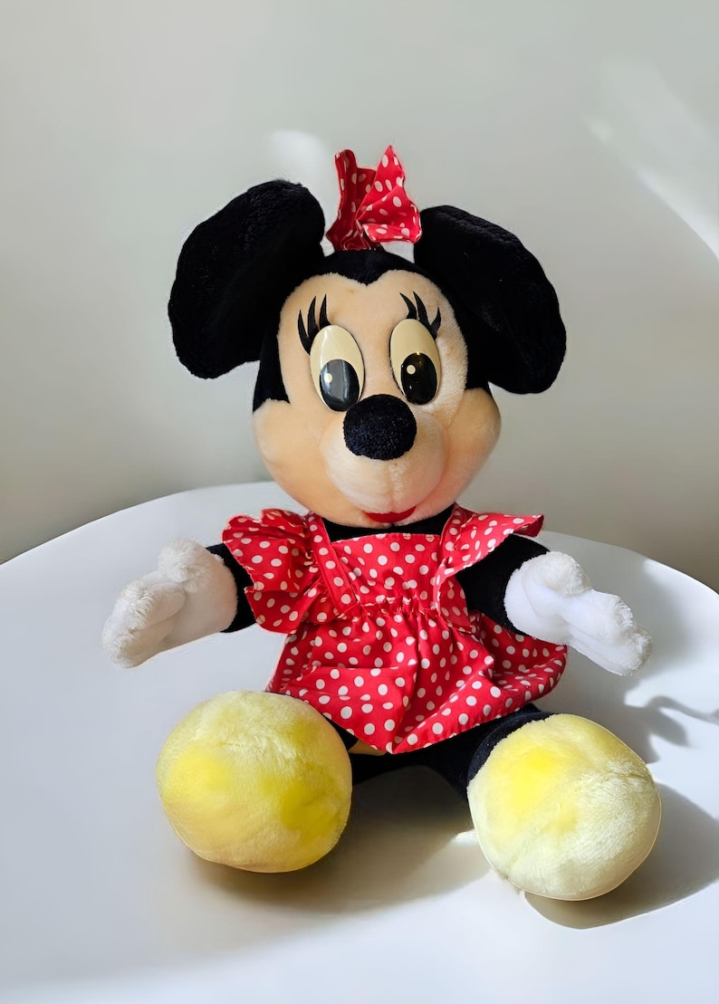 May include: A plush toy of Minnie Mouse wearing a red dress with white polka dots and a red bow in her hair. She has black ears and a black nose.
