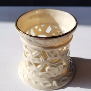 Könnte beinhalten: Ein weißer Kerzenhalter aus Keramik mit goldener Verzierung und einem filigranen Ausschnittmuster. Der Halter ist zylinderförmig mit einer breiten Basis und einer schmalen Oberseite.