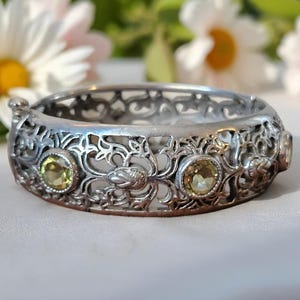 May include: Silver-toned filigree bracelet with three round, light green gemstones. The bracelet features an openwork design with floral and scroll details. The background includes white and pink flowers.