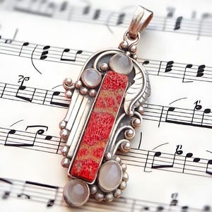 Puede incluir: Colgante de plata con una piedra central rectangular de coral rojo y tres piedras redondas más pequeñas de color claro. El colgante tiene un diseño ornamentado y detallado, sobre un fondo de notas musicales.