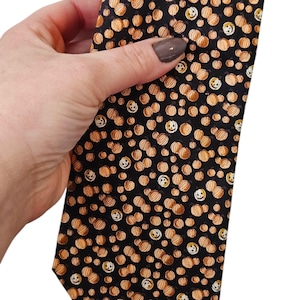 May include: A black tie with a repeating pattern of orange pumpkins with smiling faces. The pumpkins are in various sizes and are scattered across the tie. The tie is made of a smooth fabric.