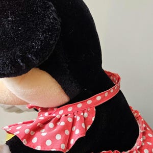 May include: A black plush toy with a red and white polka dot dress. The dress has a ruffled neckline and a white ribbon around the waist.