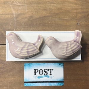 May include: Two ceramic bird figurines in a soft lavender hue, with detailed wing patterns. The birds are displayed on a white rectangular base. A business card with the word "POST" is below the birds.