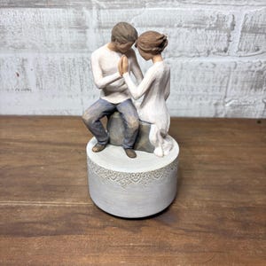 May include: A figurine depicting a couple holding hands, seated on a gray stone. The man wears a white shirt and blue jeans, while the woman is in a white dress. The base is a light gray cylinder with a decorative lace pattern.