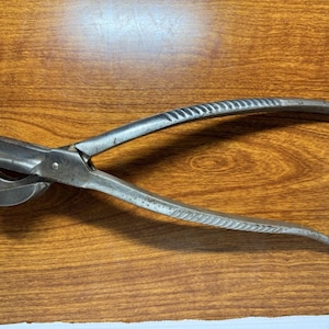 May include: A vintage, silver-colored can opener with a curved handle and a cutting mechanism. The tool is made of metal and rests on a wooden surface. The can opener is designed to open cans.