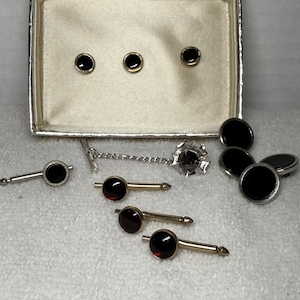 May include: A collection of men's formal wear accessories, including cufflinks, shirt studs, and a tie tack. The set features black and gold accents, with some pieces having a deep red center. The items are displayed on a white surface.