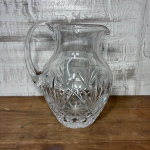 May include: A clear glass pitcher with a handle. The pitcher has a decorative, cut-glass pattern on the bottom half. The handle is curved and attached to the side of the pitcher. The pitcher is sitting on a wooden surface.