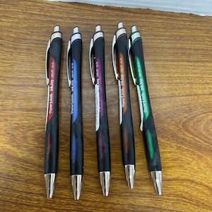 Puede incluir: Un conjunto de seis bolígrafos negros Paper Mate InkJoy 550 RT. Cada bolígrafo tiene una punta plateada y un acento de color en la parte superior, incluyendo rojo, azul, morado, naranja y verde. Los bolígrafos están colocados sobre una superficie de madera.