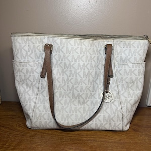 Op de afbeelding: Witte en beige tote bag met een herhaald patroon van de letters "MK". De tas heeft bruine leren handvatten en afwerking, een ritssluiting en zijvakken. Een stijlvol accessoire.