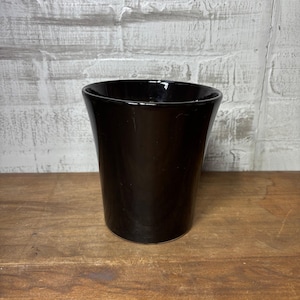May include: A black ceramic vase with a glossy finish and a flared top. The vase is cylindrical and stands on a wooden surface. The background is a white brick wall.