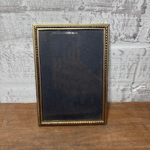May include: A rectangular gold-colored picture frame with a dark interior. The frame has a decorative, textured border. The frame is displayed on a wooden surface against a white brick wall.