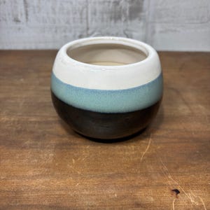 May include: A small, round ceramic pot with a white top, a teal-colored middle band, and a dark brown base. The pot is sitting on a wooden surface. The pot is likely used for holding small plants or as a decorative item.