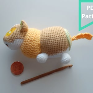 May include: Crochet pattern for a cute, beige, yellow, and white cat amigurumi toy. The pattern is available as a PDF download. The image shows the finished toy, a crochet hook, and a ball of yarn.