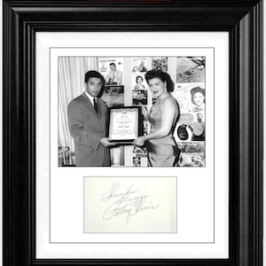 May include: A black and white framed display featuring a photograph of a man in a suit and a woman in a dress holding an award, with a collage of album covers in the background. Below the photo is a white card with the handwritten signature Patsy Cline.