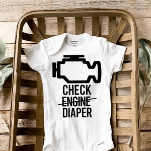 May include: A white baby grow with a black check engine light icon and the text "CHECK ENGINE DIAPER" printed on it. The grow is in a wooden basket.