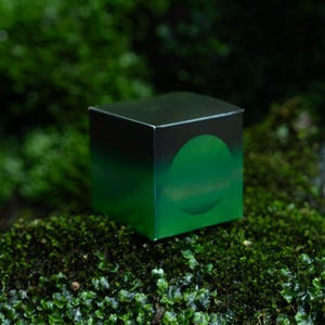 May include: A square box with a black and green gradient design and a green circle on the side. The box is placed on green moss. The box has a modern, minimalist aesthetic.