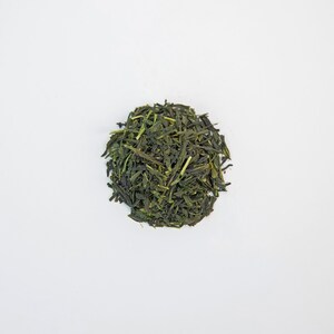 May include: A circular pile of dried green tea leaves on a white backdrop. The tea leaves are a deep green hue, with a slightly textured appearance. The leaves are small and thin, with some stems visible.