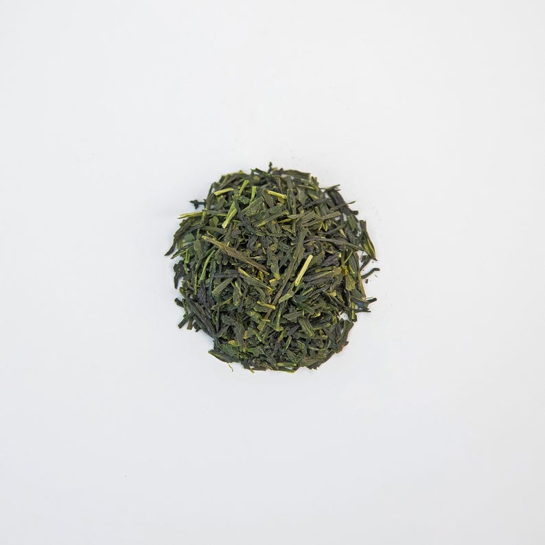 May include: A circular pile of dried green tea leaves on a white backdrop. The tea leaves are a deep green hue, with a slightly textured appearance. The leaves are small and thin, with some stems visible.