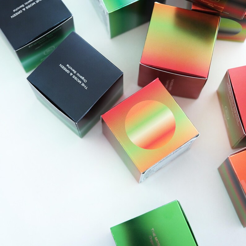 May include: An assortment of square boxes with gradient colour schemes of green, red, and gold are arranged on a white surface. Some boxes are black with green accents and text that reads "THE MOSS & GREEN Organic Sencha".