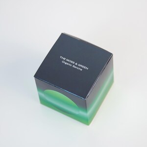 May include: A square box with a gradient design, transitioning from dark blue to green. The text "THE MOSS & GREEN Organic Sencha" is printed on the top. The box is likely packaging for a tea product.