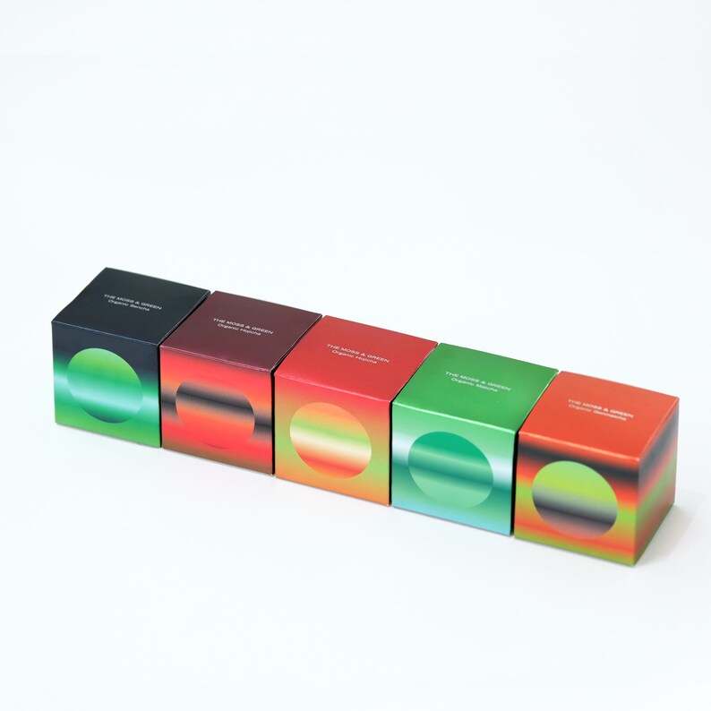 May include: Five square boxes in a row, each with a different colour gradient and a circular design. The boxes are black, burgundy, red, green, and orange. The text "THE MORE & GREEN Organic Beauty" is printed on the top of each box.