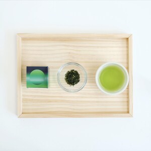 May include: A wooden tray displays a small box with a green gradient design, a small glass bowl of dried green tea leaves, and a white teacup filled with green tea. The items are arranged on a white surface.